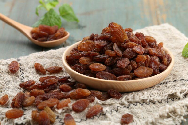 A shallow wooden dish with dried raisins, with more raisins scattered on the table