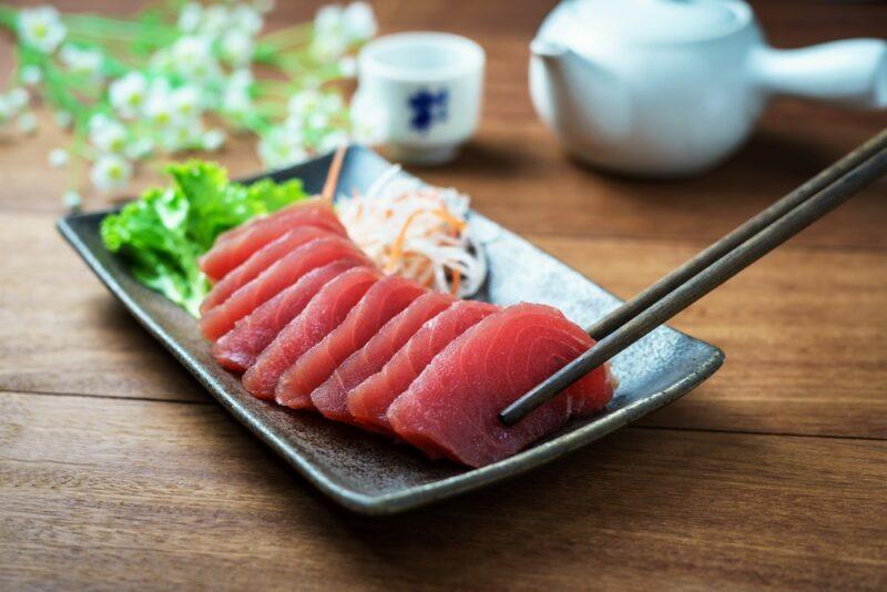 A wooden table with a black dish of raw sliced tuna, with a piece being grabbed by chopsticks