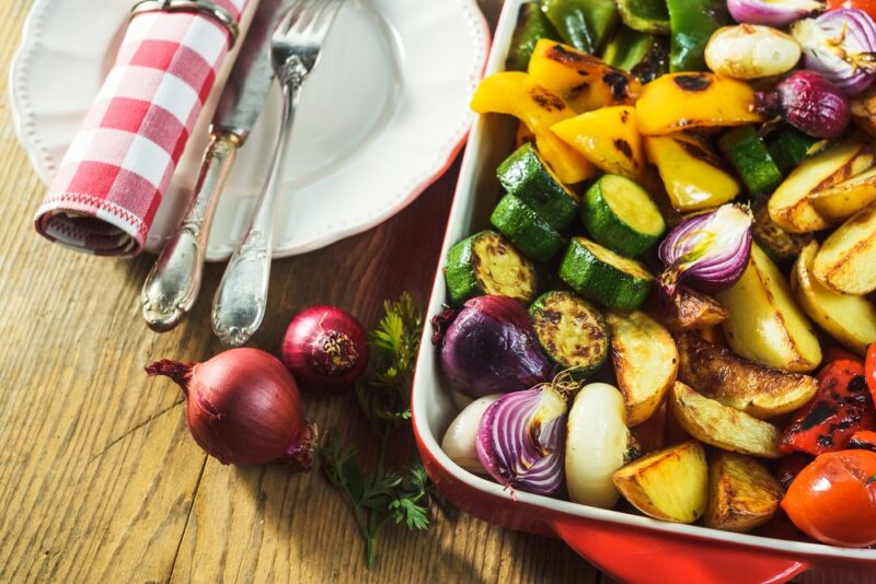 An oven dish containing many types of roast vegetables, next to a white plate with a fork and a knife