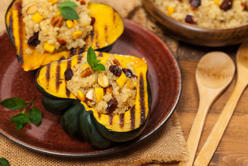 A brown-red dish containing an acorn squash that has been sliced in half, roasted, and stuffed, next to two spoons and a bowl of the stuffing