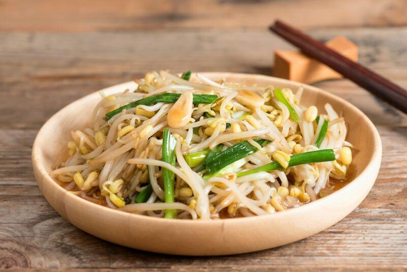 A shallow light brown dish containing stir fried bean sprouts and other vegetables, next to a pair of chopsticks.