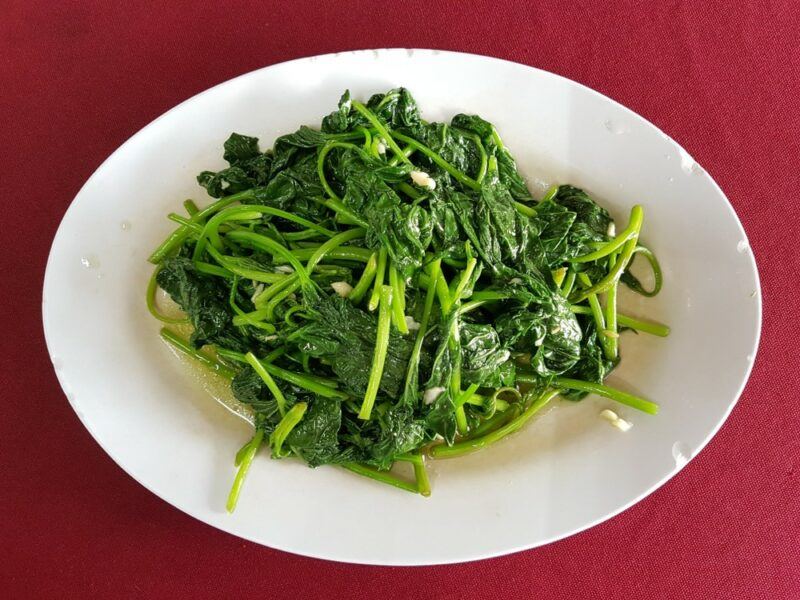 A red table with a white plate that contains a dish of stir fried sweet potato leaves. This could be a delicious side for a Chinese meal.