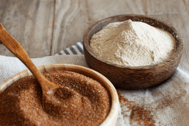 A wooden table with a bowl of teff flour and a bowl of brown teff grains with a spoon.