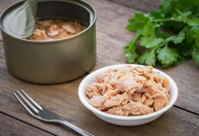 A small white bowl of tuna, next to a can of tuna on a wooden table