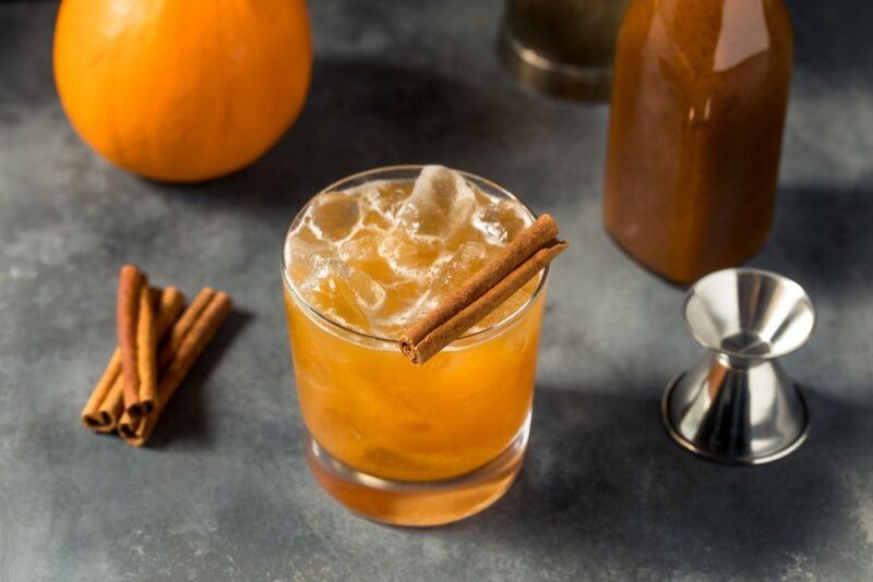 An old fashioned glass with a drambule cocktail, next to cinnamon sticks, an orange, and a cocktail jigger