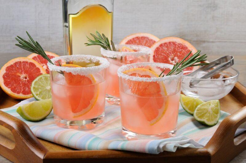 Three glasses of a paloma cocktail, each with salt around the rim and garnished with a sprig of rosemary, with a bottle and cut pieces of grapefruit and limes in the background.