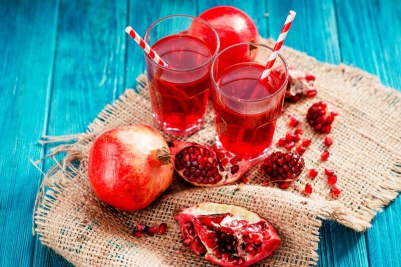 Two glasses of pomegranate juice with straws, with some pomegranates on some type of sack or brown cloth