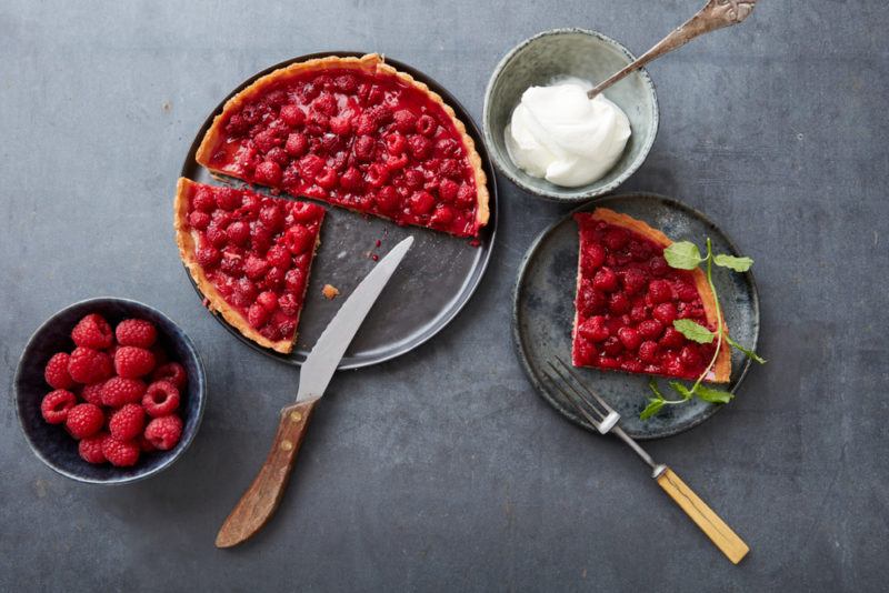 A dish of fresh raspberry and apple pie, next to a plate with a piece of the pie, a bowl of raspberries, and a bowl of sugar