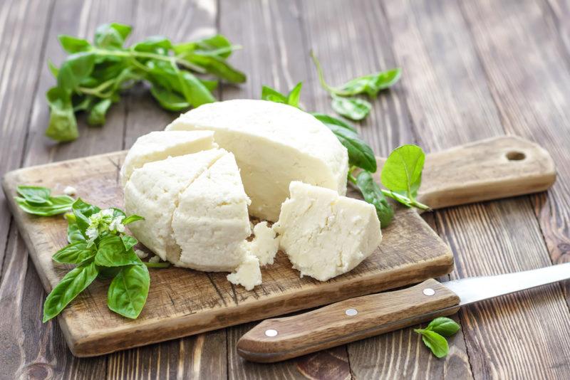 A wooden board with soft cheese that has been cut into wedges, fresh herbs, and a knife