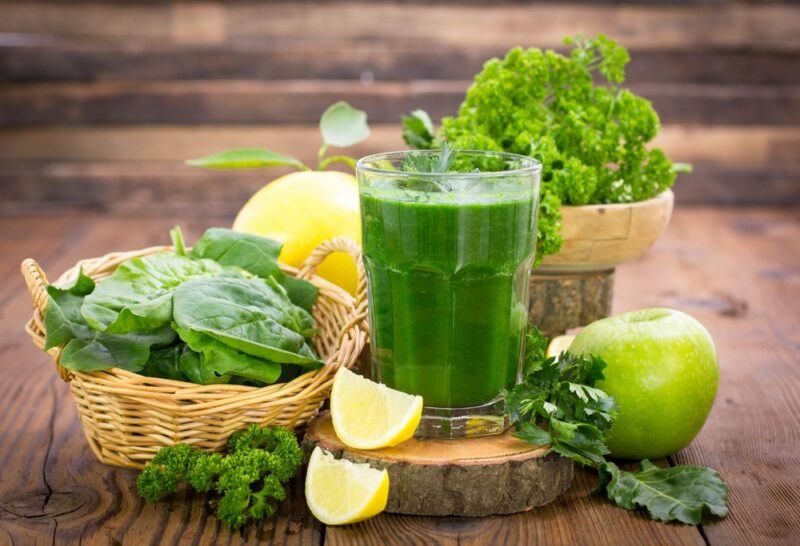 A table with a glass containing a vegetable smoothie, surrounded by fresh vegetables, an apple, and a grapefruit