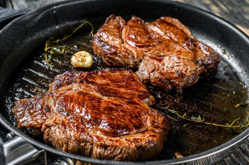 A black fry pan with two chuck eye steaks that are being cooked next to some rosemary and garlic