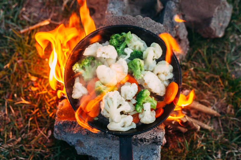 A skillet over a campfire with broccoli, cauliflower, and carrots