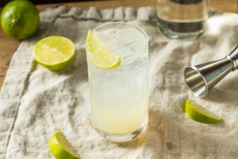 A cloth with a single tall glass containing a gin rickey cocktail, with various limes scattered around.