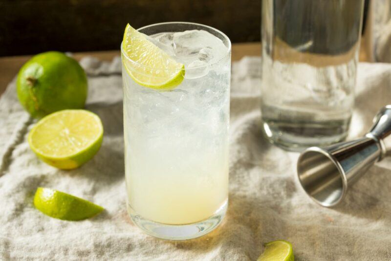 A tall glass containing a gin rickey cocktail with ice, next to lime pieces and a cocktail jigger