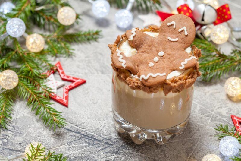 A large glass containing a gingerbread cocktail made with gingerbread vodka, where there is part of a Christmas tree on the table as well