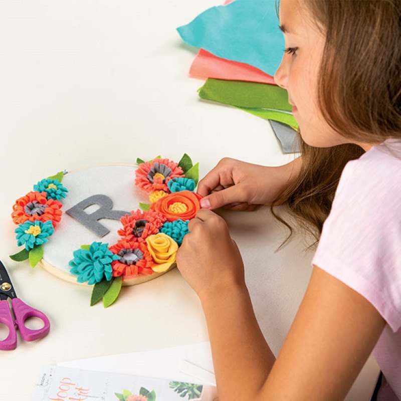 A girl at a table working on a craft attachinh fabric flowers to a circle with a felt R in the middle 