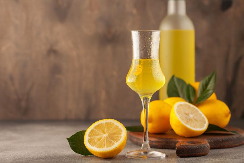 A glass of Limoncello in the foreground and a bottle of Limoncello in the background, with sliced lemons on the table