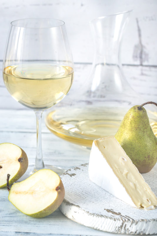 A glass and a carafe of savignong blanc wine on a white table, next to a whole pear, two pear halves, and a wedge of camembert