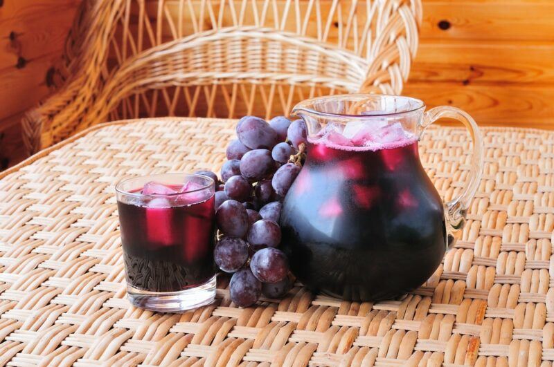 A wicker chair next to a wicker table that has a glass of grape juice, a jug of the juice, and some grapes