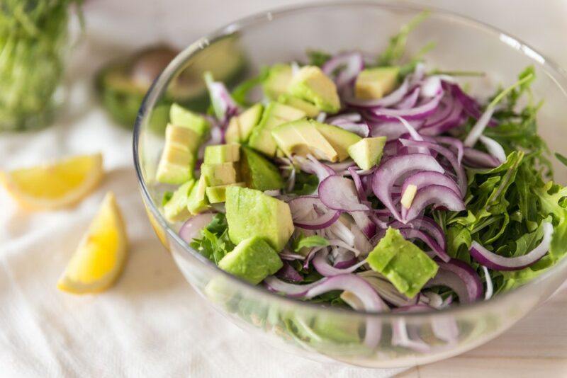 A glass bowl containing a avocado and red onion salad, on a white cloth with some lemon wedges