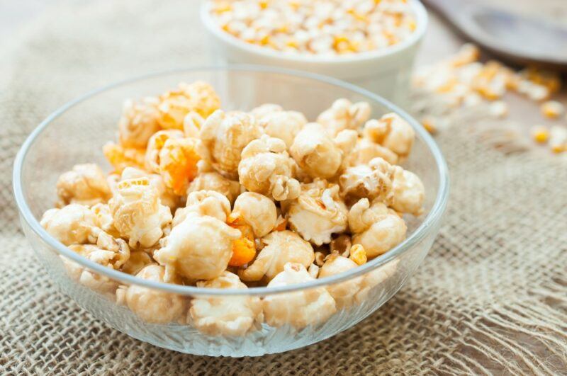 A glass bowl containing caramel and white chocolate popcorn, with popcorn kernels in the background