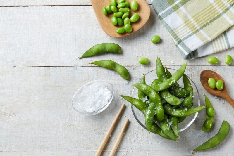 A table with a selection of dishes and other items, including a glass bowl of edamame, some salt, chopsticks and a tea towel