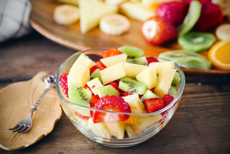 A glass bowl containing a fresh fruit salad, with more fresh fruit in the background