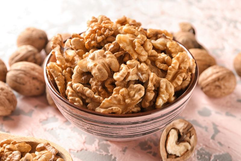 A ceramic or glass bowl that contains shelled walnuts, with whole walnuts on the table