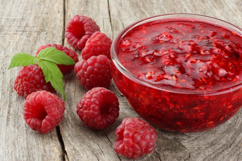 A glass bowl of raspberry sauce next to some fresh raspberries