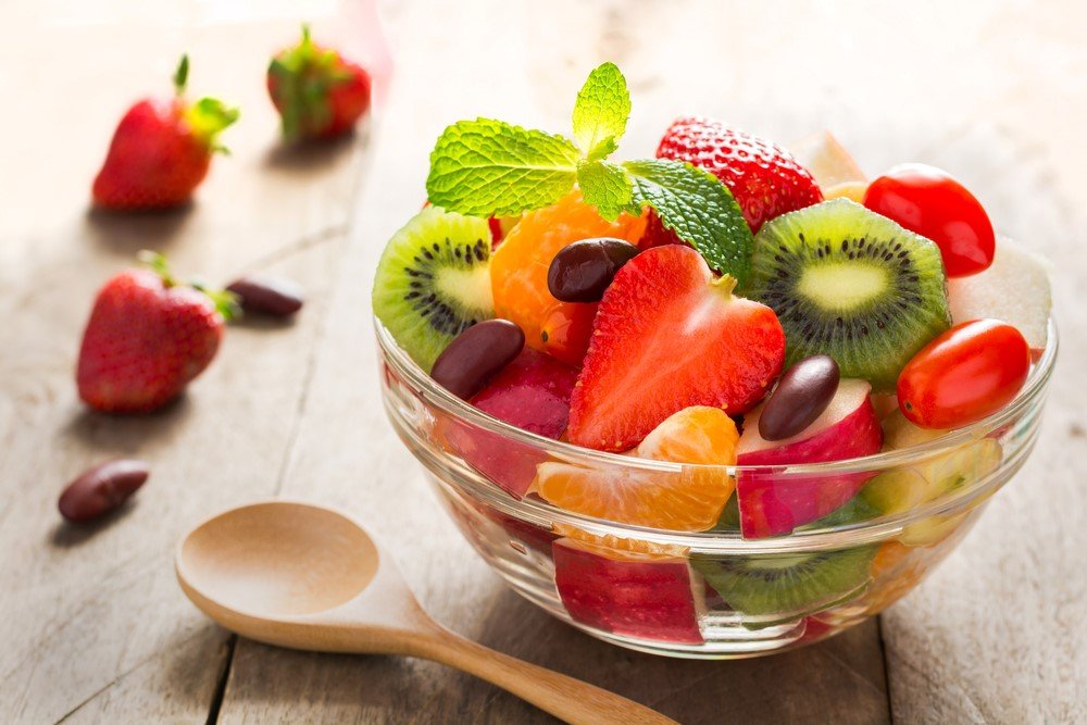 A glass bowl of fruit salad, next to a wooden spoon and some berries on a wooden table