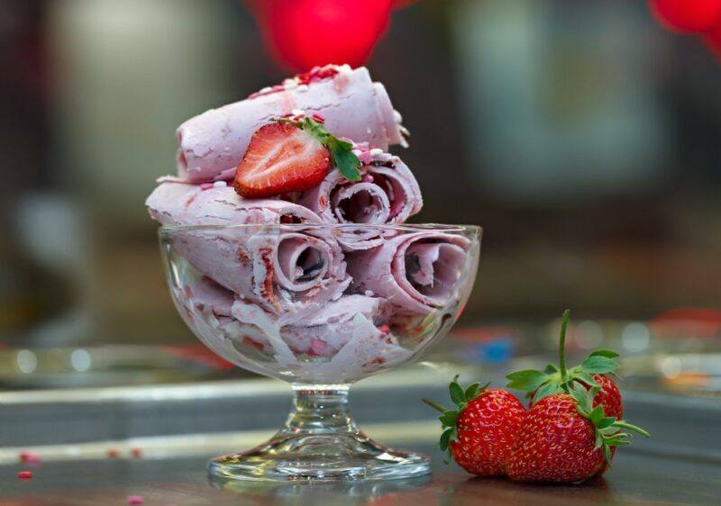 A glass bowl containing purple rolled ice cream with a strawberry, plus more strawberries on a table