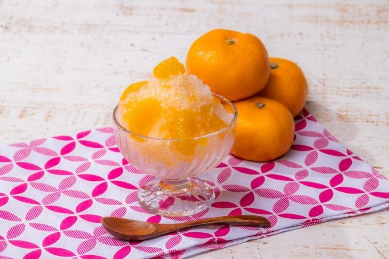 A pile of oranges or mandarins, next to a bowl of orange shaved ice and a spoon