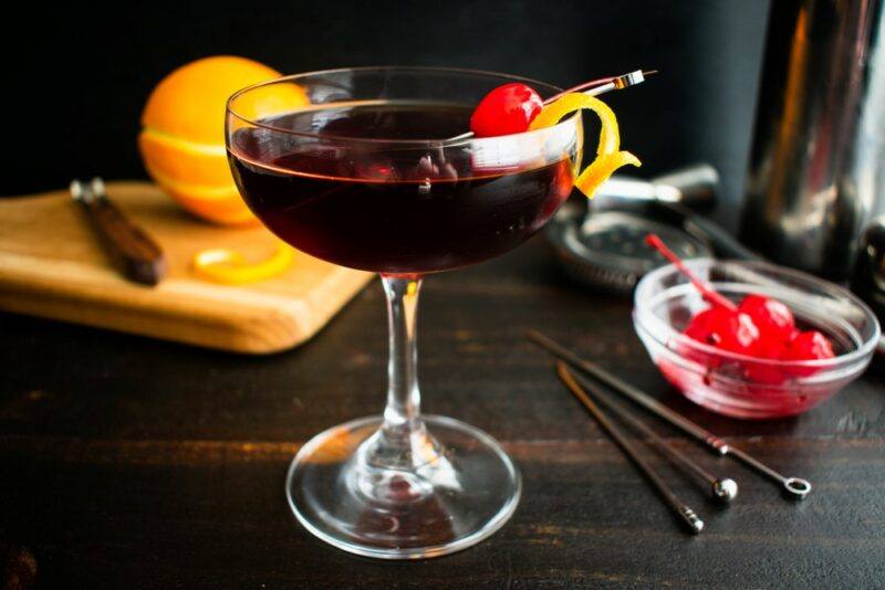 A coupe glass that contains a Reese's Manhattan cocktail, garnished with an orange twist and a cherry, next to stirrers, an orange, and a small dish of cherries