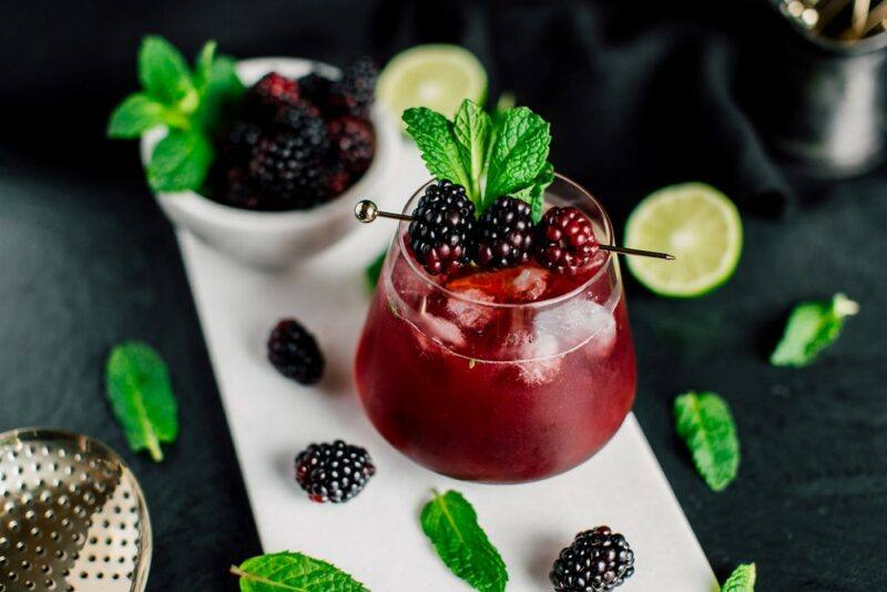 A glass of a cocktail called bee sage on a white plate, surrounded by blackberries and sage leaves