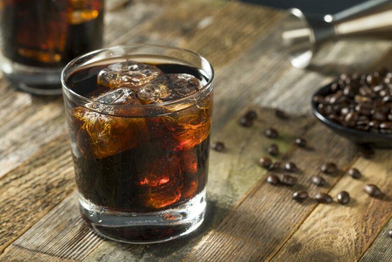 Two glasses of a black Russian cocktail on a wooden table surrounded by coffee beans