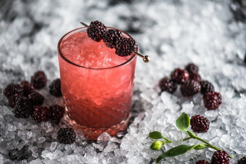 A single glass containing a blackberry moonshine margarita cocktail, surrounded by a bed of crushed ice and plenty of blackberries