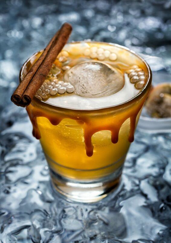 A glass with a ball of ice, a cinnamon stick, and caramel sauce, containing a butterscotch gold dessert martini