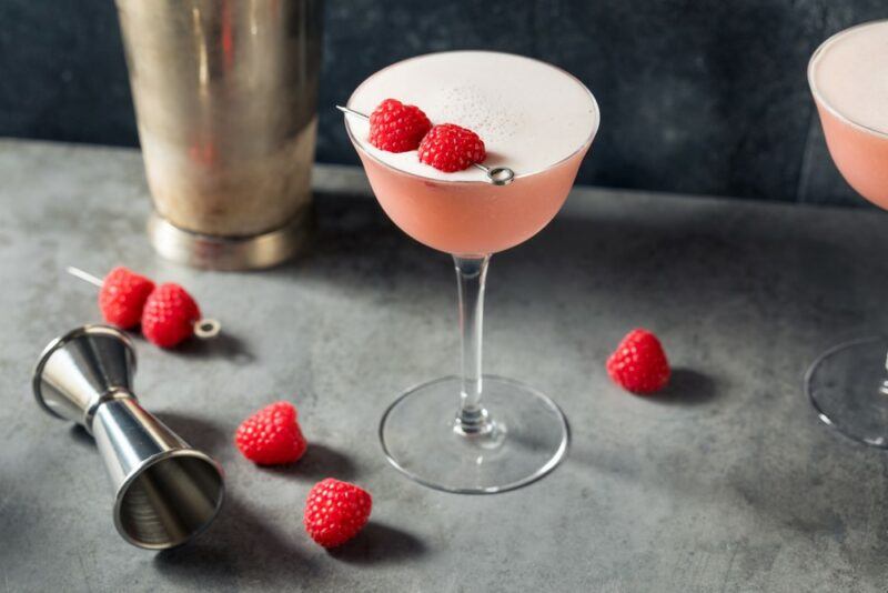 A light pink clover club cocktail in a coupe glass with raspberries as a garnish and on the table