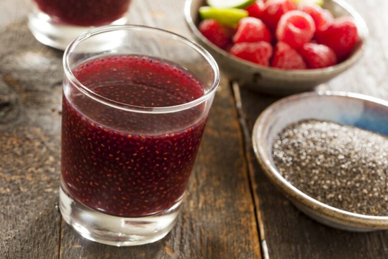 A glass containing a raspberry chia drink, next to bowls of raspberries and chia seeds