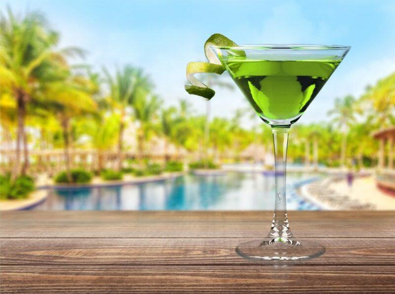 A martini glass on a table containing a green tea of wisdom cocktail, overlooking a large pool that's surrounded by many trees