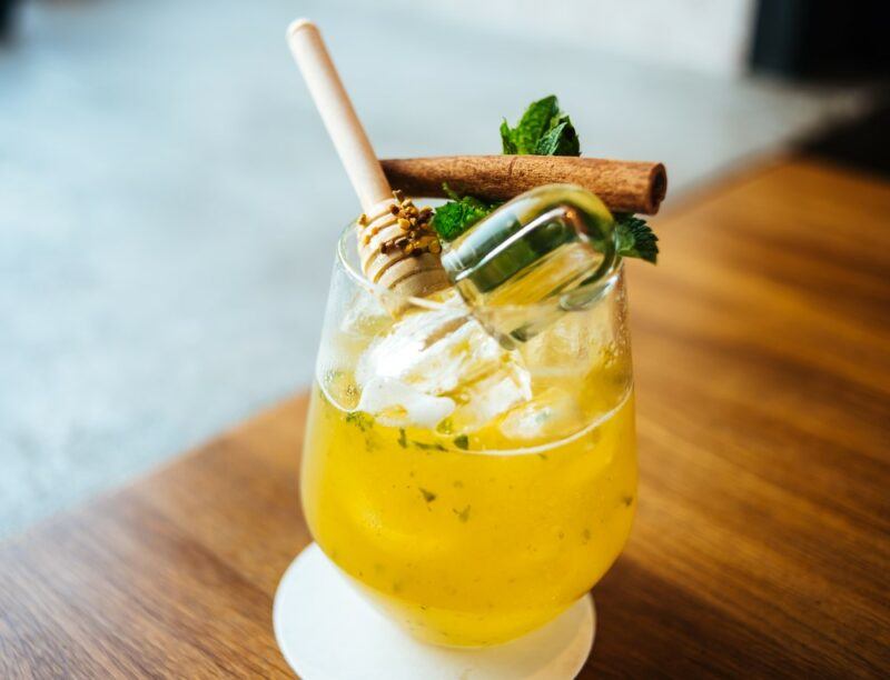 A glass containing a tractor fuel cocktail with a honey stirrer and cinnamon stick as a garnish