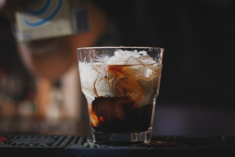 A glass containing a white Russian cocktail on a bar with an out of focus background