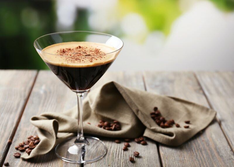 A glass containing an espresso martini on a wooden table with a cloth containing coffee beans