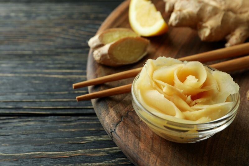 A dark board with fermented ginger slices, next to chopsticks and ginger root