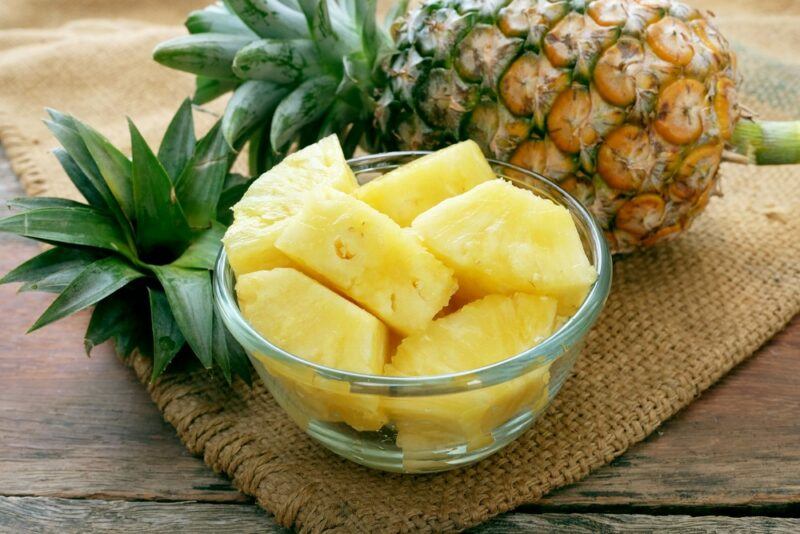 A glass bowl of pineapple pieces, next to the top of a pineapple and a whole fresh pineapple.