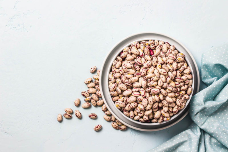 A glass jar of beans on a light background next to a blue cloth, with some of the beans on the table