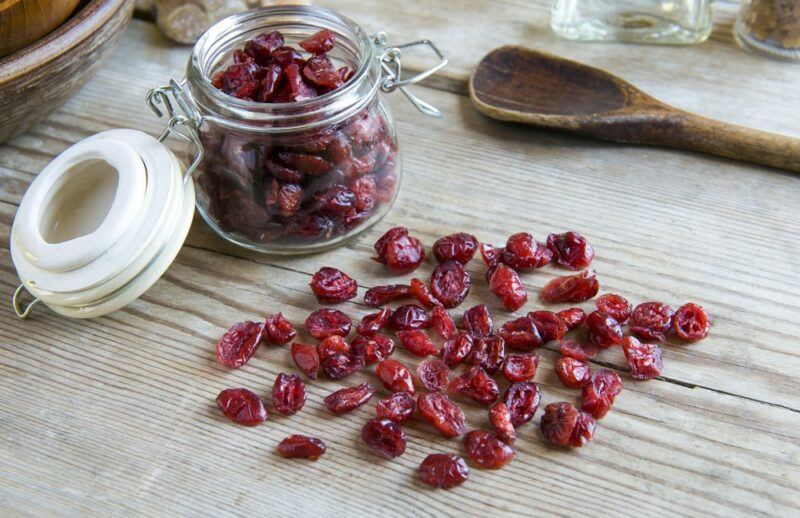 A jar of dried cranberries on a table, next to a wooden spoon, with many dried cranberries in front of it