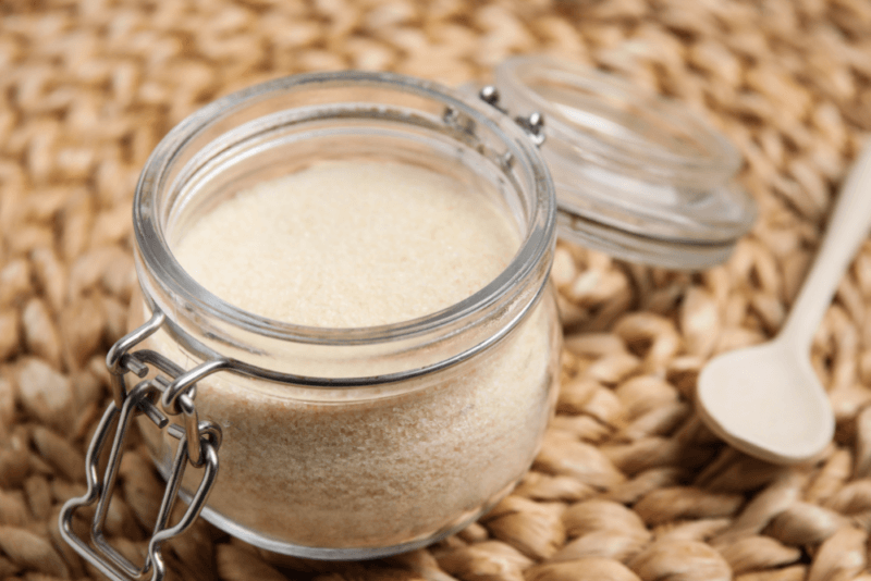 A glass jar containing gelatin powder next to a small wooden spoon of the powder