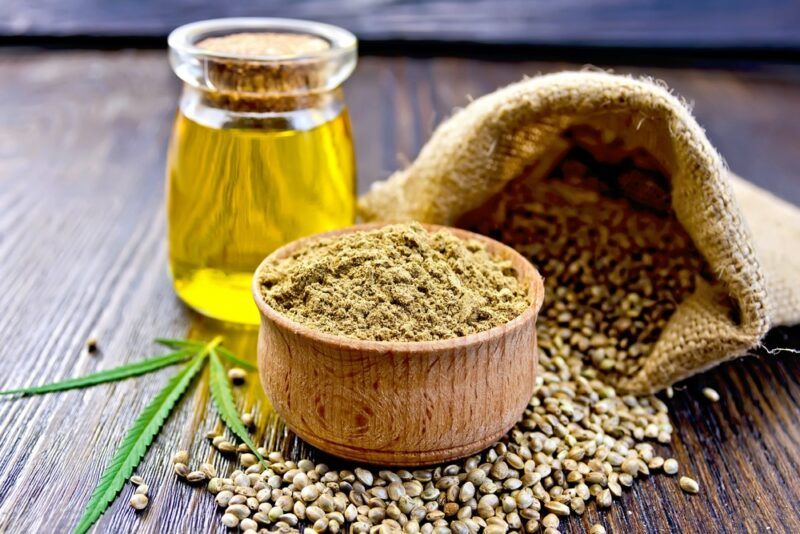 A wooden table with a bag of hemp seeds, a dish of hemp powder, and a glass jar of hemp oil