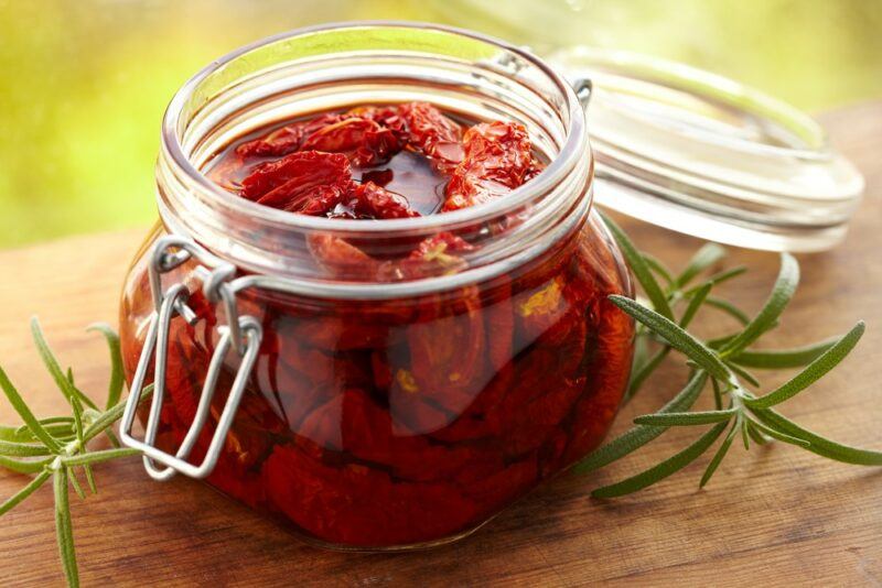 A small glass jar next to some sprigs of herbs, packed full of sundried tomatos